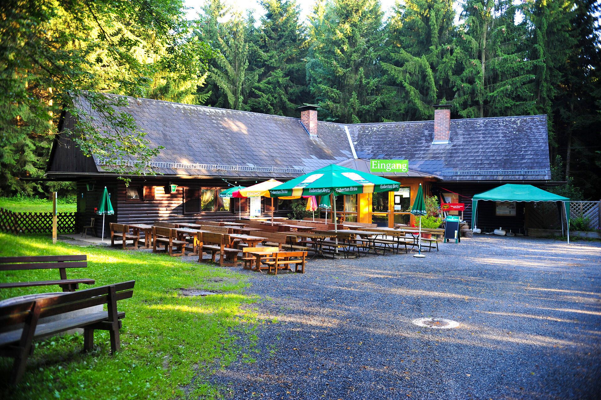 Umgeben von üppigem Grün und hohen Bäumen lädt die einladende Hütte im Naturpark Geras dazu ein, eine wohlverdiente Pause einzulegen. Die rustikalen Holztische und bunten Sonnenschirme schaffen eine gemütliche Atmosphäre, perfekt für gesellige Stunden in der Natur.